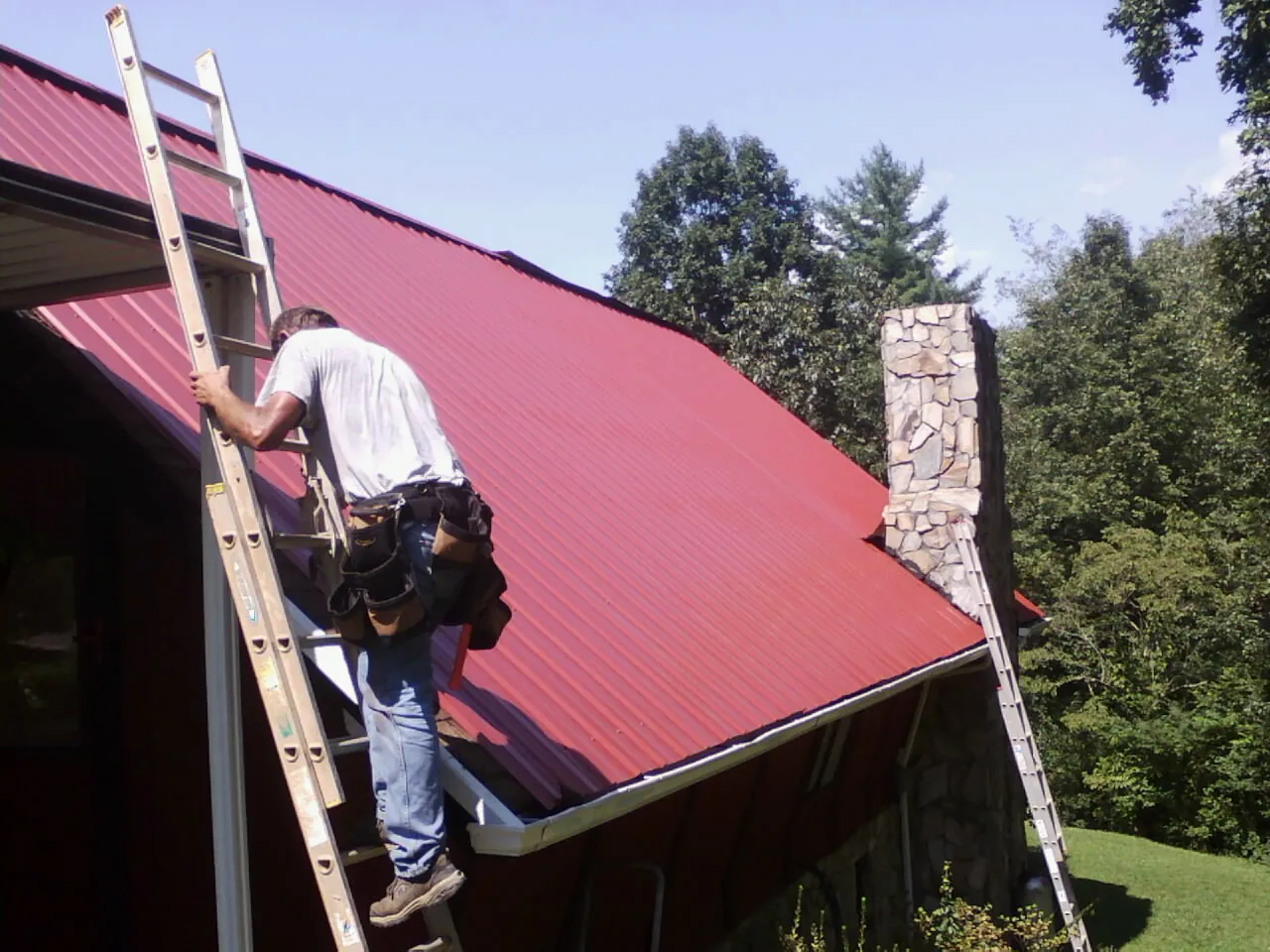 Roofer on a ladder beside a red metal roof and stone chimney — Soffit & Fascia Repair by Roofing Brothers in New Providence